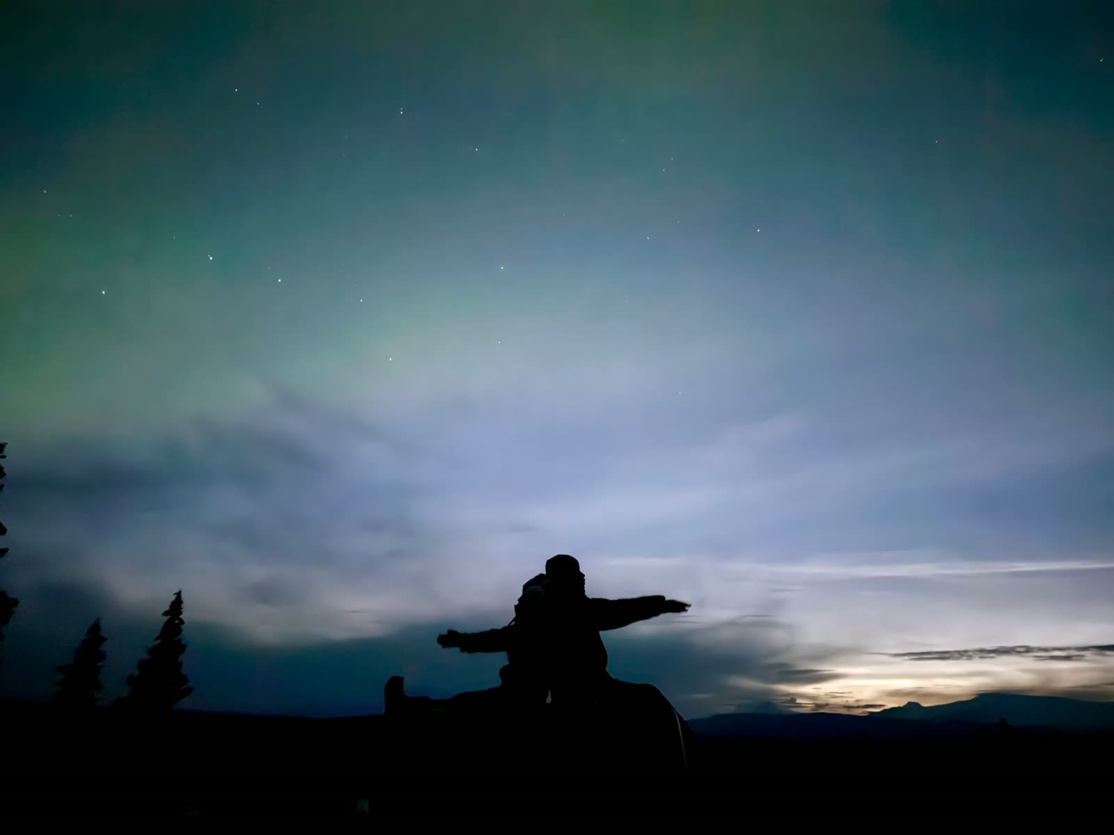 A silhouette with arms outstretched beneath a wide green aurora and distant hills