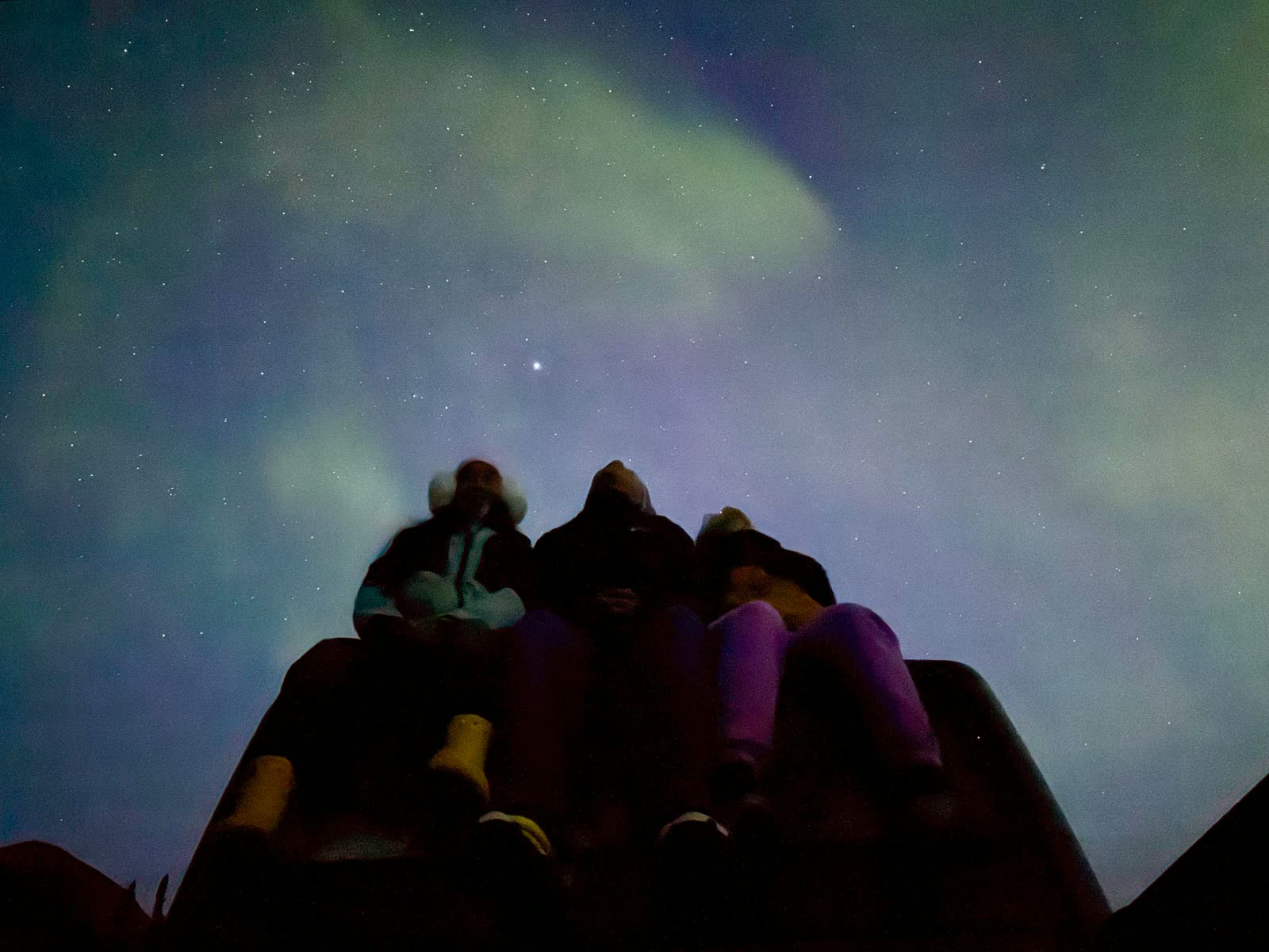 Three silhouettes seated on a rock watching the aurora and a bright star