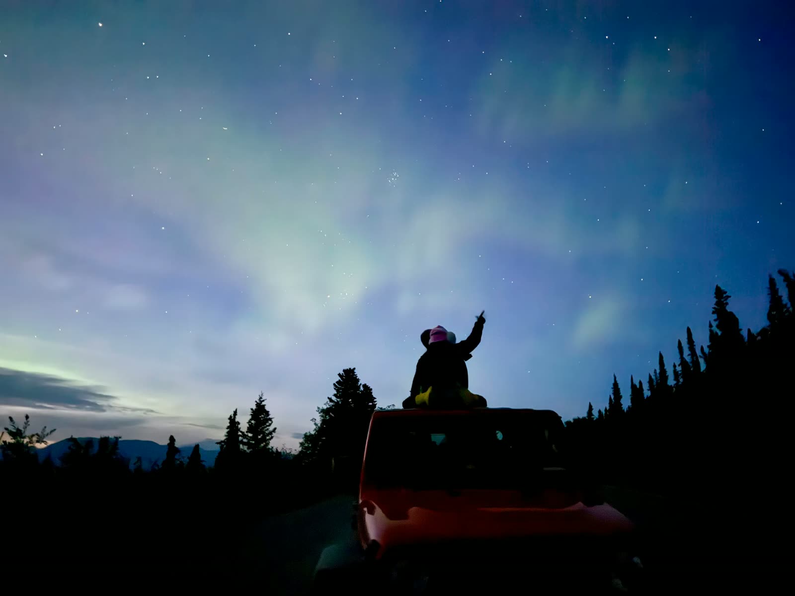 A child on a truck roof pointing up at the aurora above the spruce line
